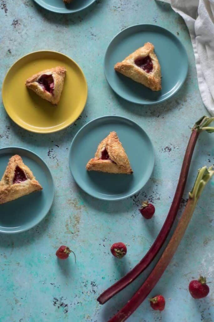 strawberry rhubarb turnovers on blue and yellow plates on a blue surface with strawberries and rhubarb next to them.