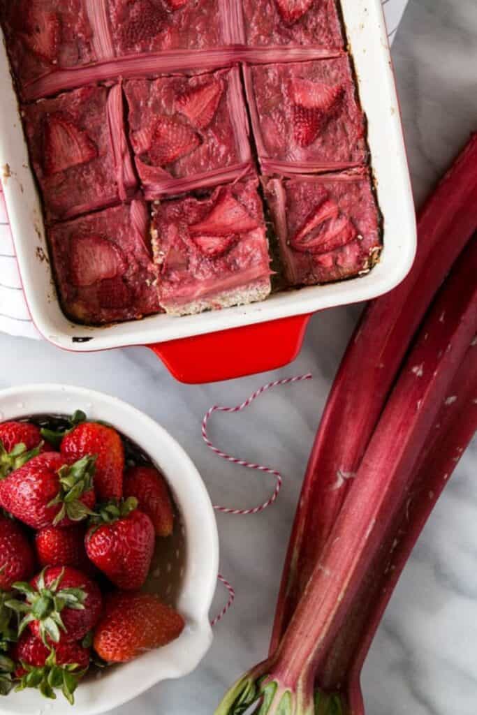 strawberry rhubarb oat bars in a white dish with red handles sittin on a white and blue striped towel on a marble surface with a bowl of strawberries and rhubarb next to it.