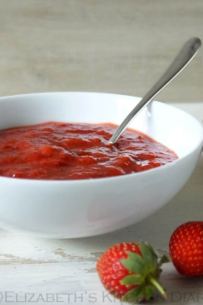 Strawberry rhubarb compote in a white bowl with a silver spoon and strawberries laying on the wood surface next to the bowl.