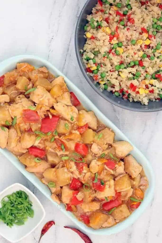 Pineapple chicken on a white rectangular plate on a marble counter and a bowl of rice and veggies.