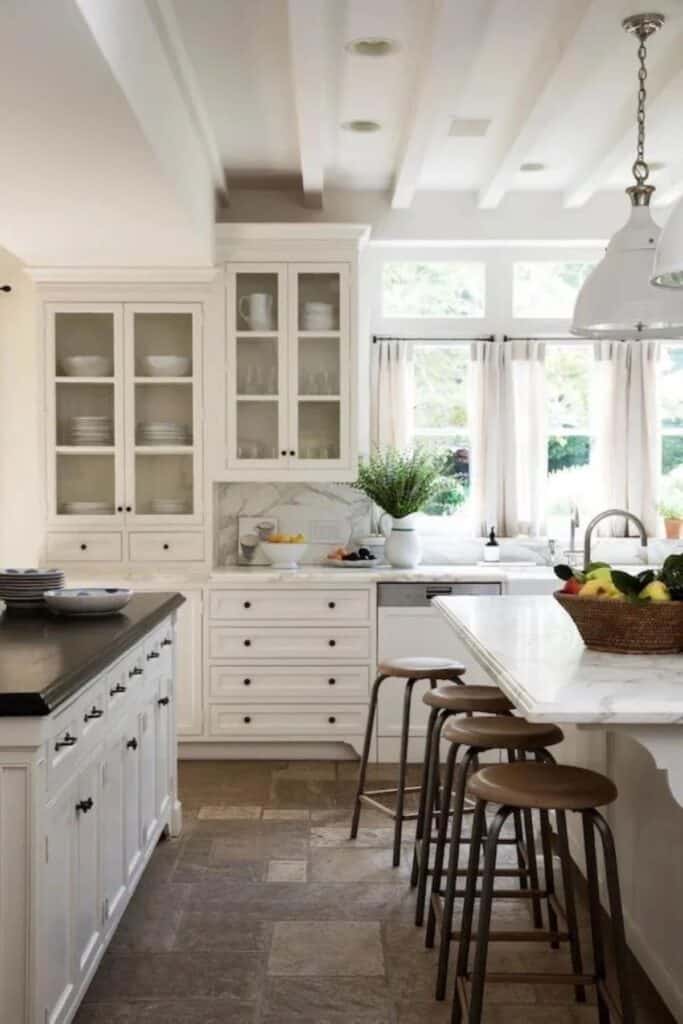A kitchen with lots of windows, cream cabinets with black hardware and the uppers have glass fronts so you can see the dishes inside.