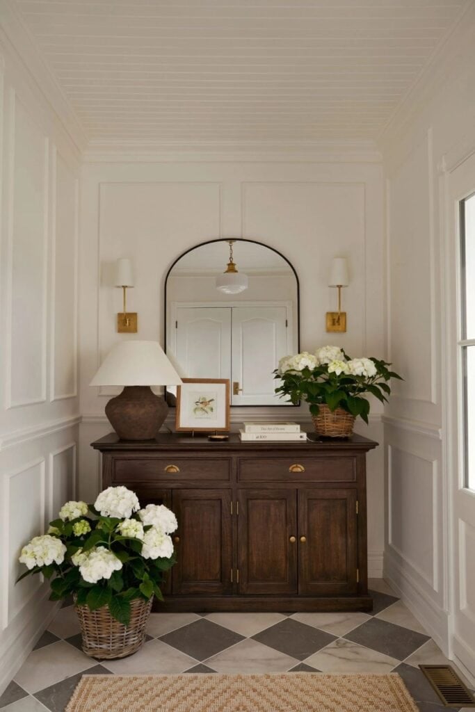 A dresser that holds a lamp and a potted hydranges with a framed artwork and books with a mirror and scones on the wall. There is aother hydranges on the checkered tile floor.