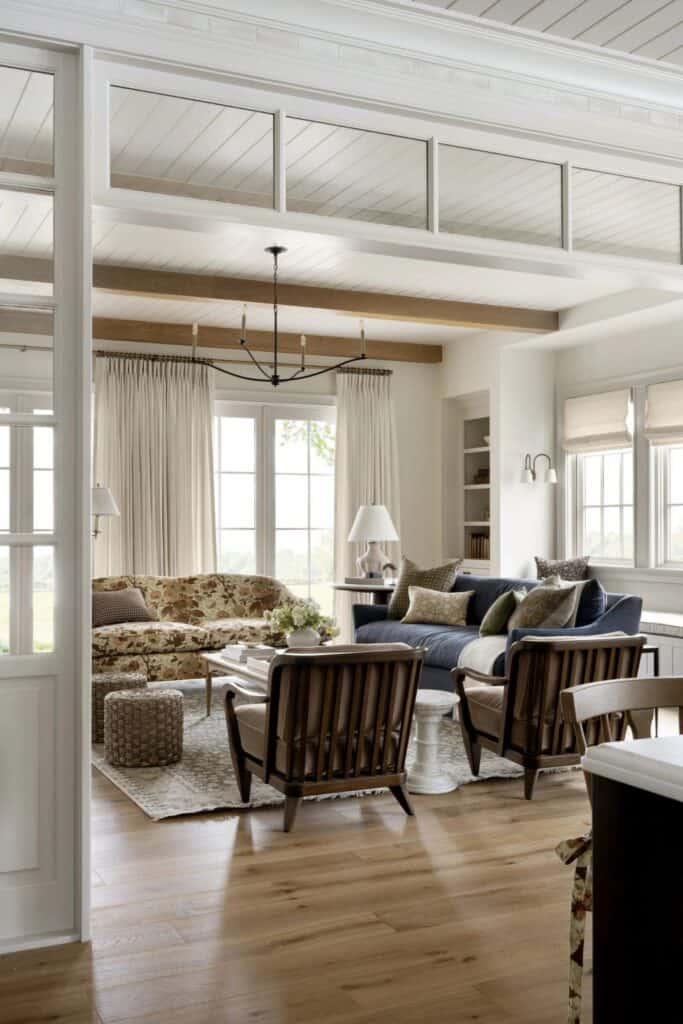 A living room with lots of windows and linen drapes with two sofas and two chairs. There is a large chandelier, lamps and wall sconces as well.