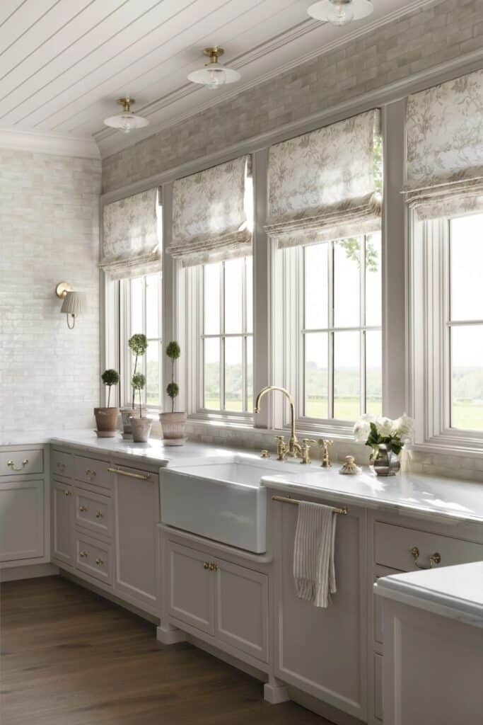 A kitchen with tons of windows, light gray counters with a farmhouse sink, terracotta pots with topiaries and a wall of tile with a sconce.