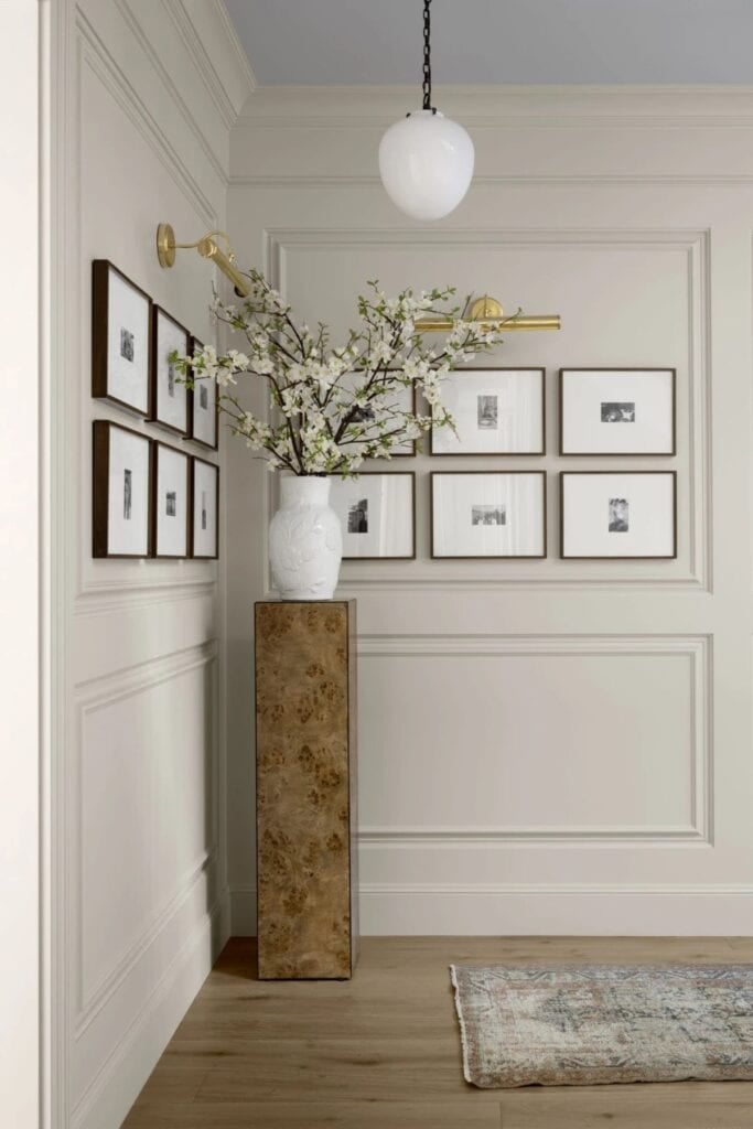 A wall with picture molding and a gallery wall of family phots with two wall sconces above and a pedestal table with a vase of flowers.