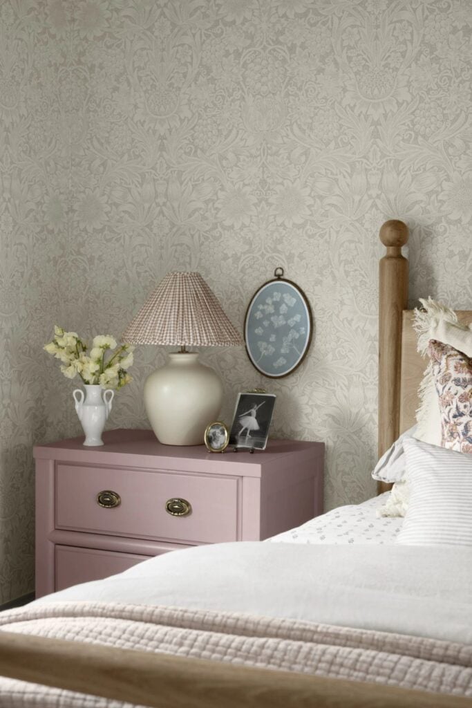 a bedroom with wallpaper, a pink side table holding a lamp, flowers, and two framed phots.