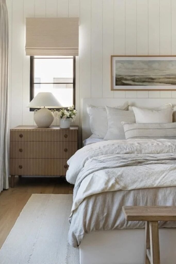 a bedroom with neutral cozy bedding with lots of throw pillows, a wood bench at the end and a wood nightstand. The walls are vertical shiplap and a landscape piece of artwork above the headboard.
