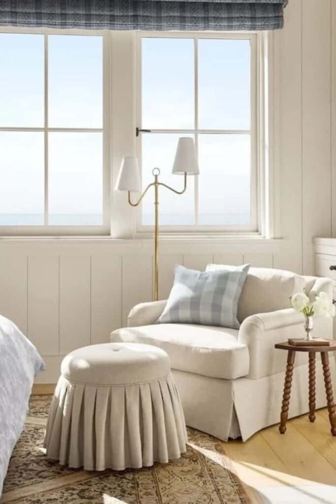 A sitting chair and ottoman that are skirted, a wooden side table with turned legs and a brass lamp next to it with lots of windows.