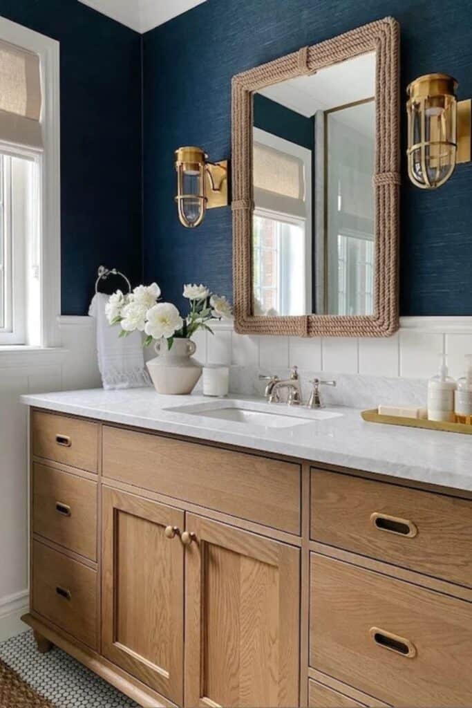 A bathroom with a wood cabinet and the lower walls are white vertical shiplap with the upper being a navy grasscloth. The mirror frame is nautical rope and the wall sconces are nautical inspired as well.