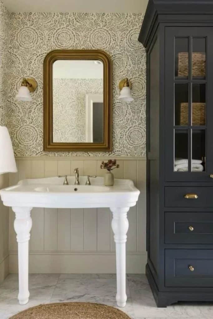 A bahtroom with a white pedestal sink next to a large navy piece of cabinetry with glass fronts. The lower part of the walls are vertical shiplap and the upper walls is wallpaper.
