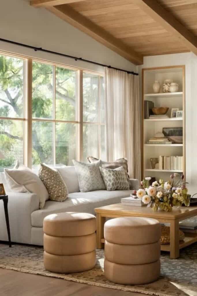 A living room with tons of windows, a sofa with 5 throw pillows, a wooden coffee table holding accessories and a vase of flowers and built--ins holding books and accessories.