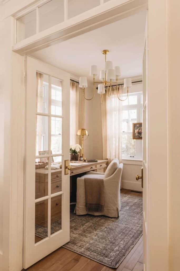 the view into a room that is an office with a wood desk, an upholstered chair, a chandelier and tons of windows.