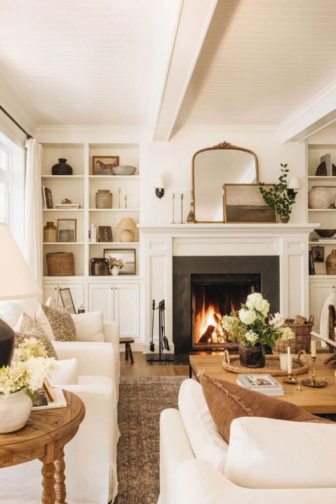 a family room with a fire going in the fireplace, a large mirror and a landscape print on the mantel flanked by two built-ins and a counch, two chairs and a coffee table.